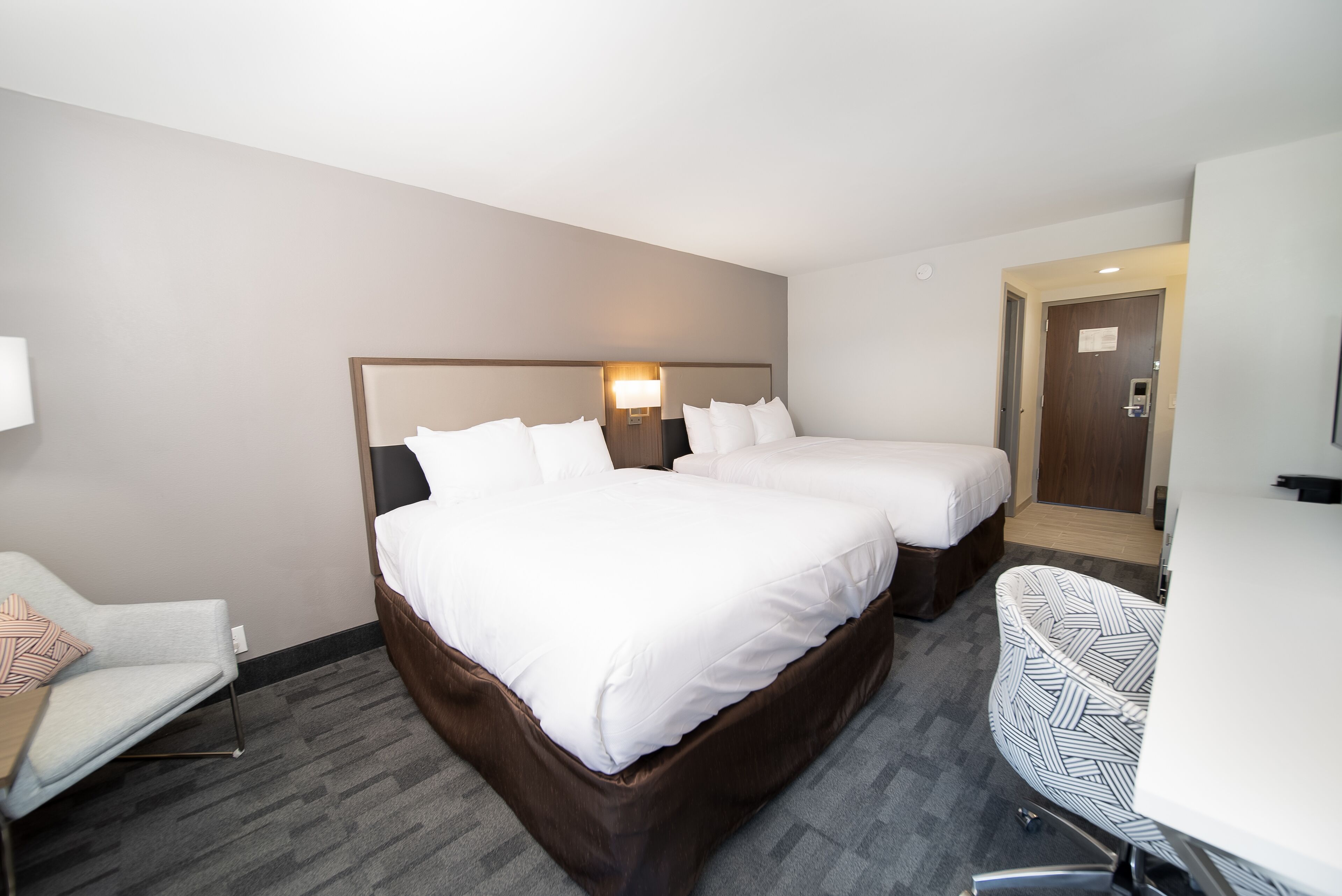 A hotel room with two beds, a desk, a chair, and a lamp.