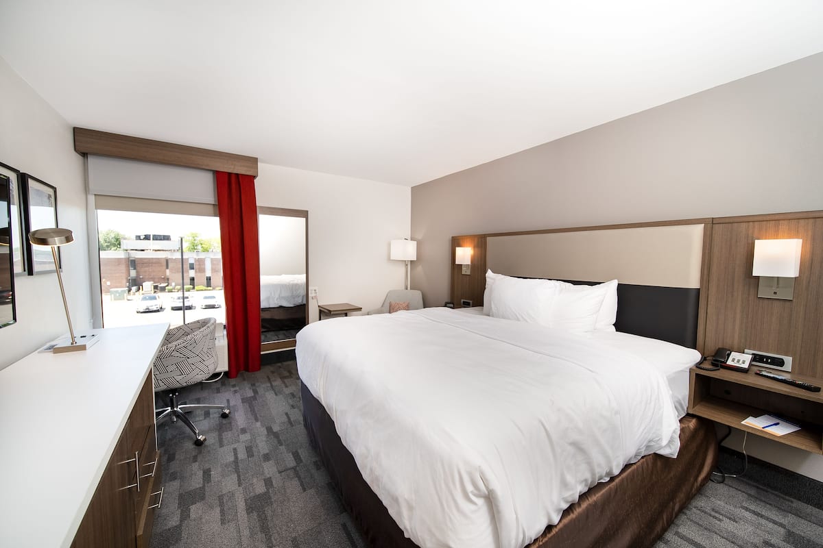 A hotel room with a large bed, a desk, a chair, a lamp, and a view of the outside.