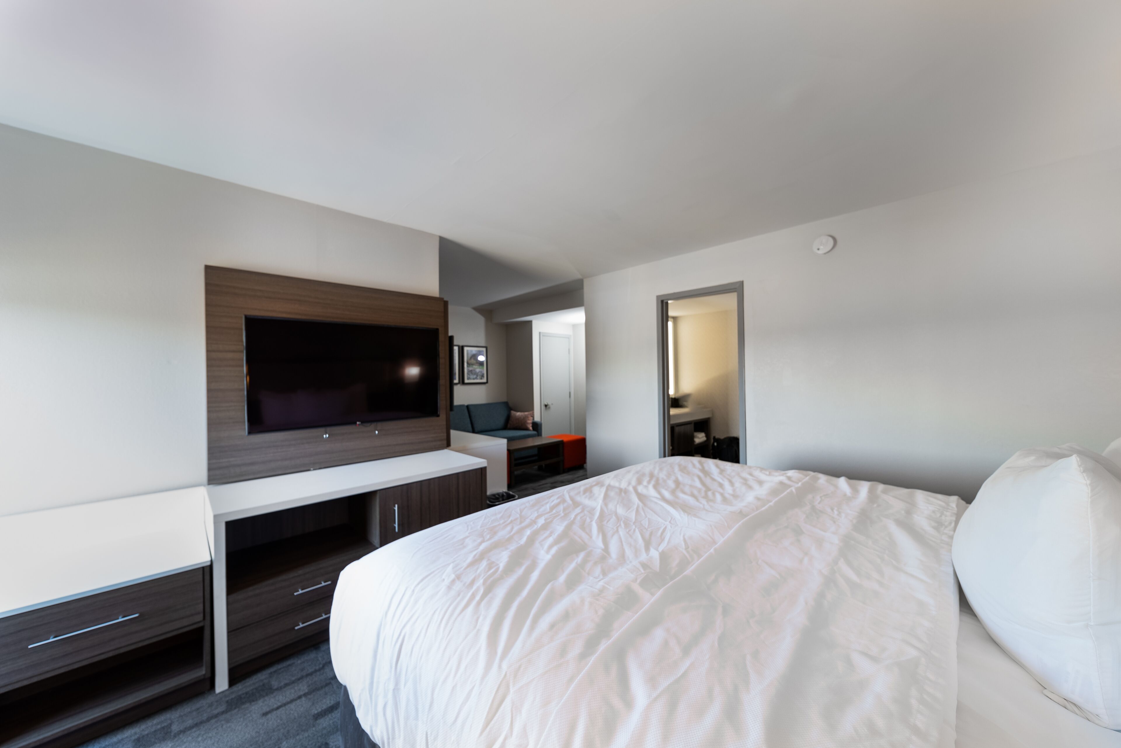 Une chambre d’hôtel moderne dotée d’un grand lit, d’un téléviseur à écran plat fixé sur un panneau en bois, d’un bureau avec des tiroirs et d’un petit coin salon comprenant une chaise et une table.