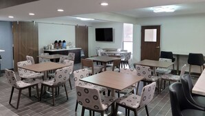 A modern dining area with multiple tables and chairs, a kitchen counter, and a television on the wall.