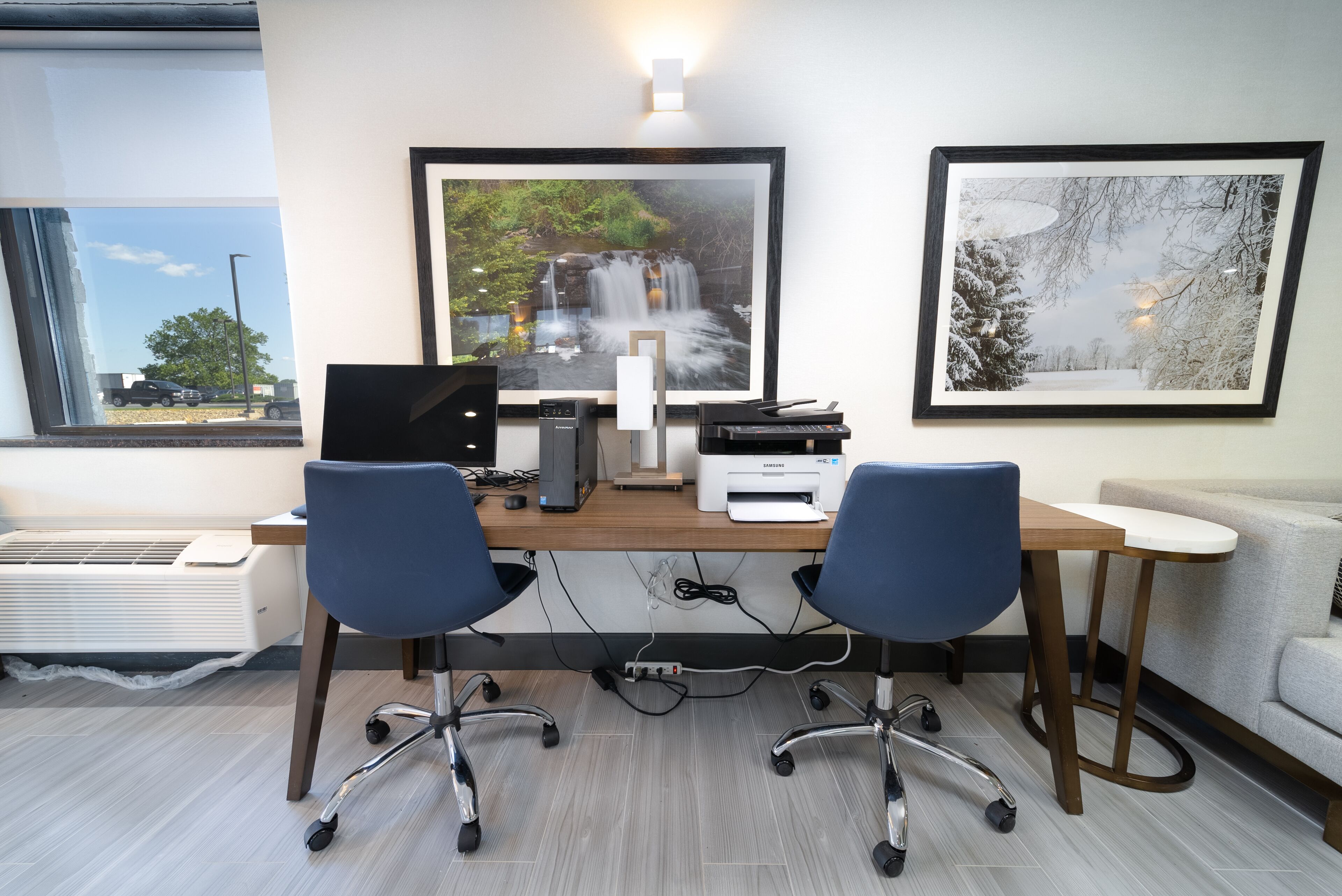 A modern office with two blue office chairs, a wooden desk, a computer, a printer, and framed pictures on the wall.