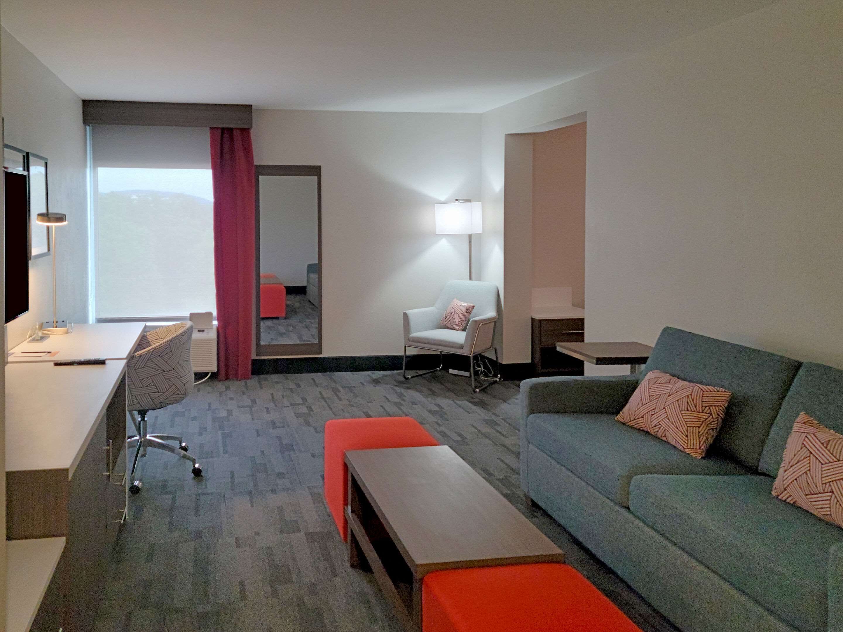 A modern hotel room with a desk, a grey sofa, a red ottoman, a white armchair, and a large mirror.