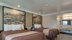 Une chambre d’hôtel avec deux lits, un grand tableau au mur, une salle de bain avec un miroir et un lavabo, et un plancher en bois.