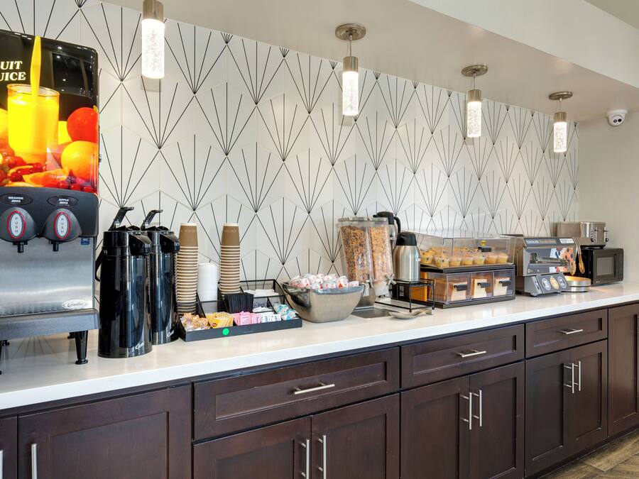 A self-service breakfast station with a fruit juice dispenser, coffee makers, and various food items.