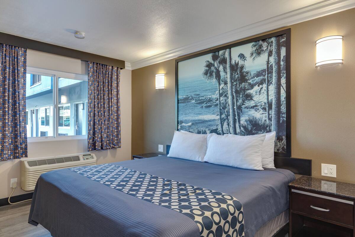 Ein Hotelzimmer mit einem großen Bett, einem Nachttisch, einer Wandlampe und einem großen Gemälde einer Strandansicht.