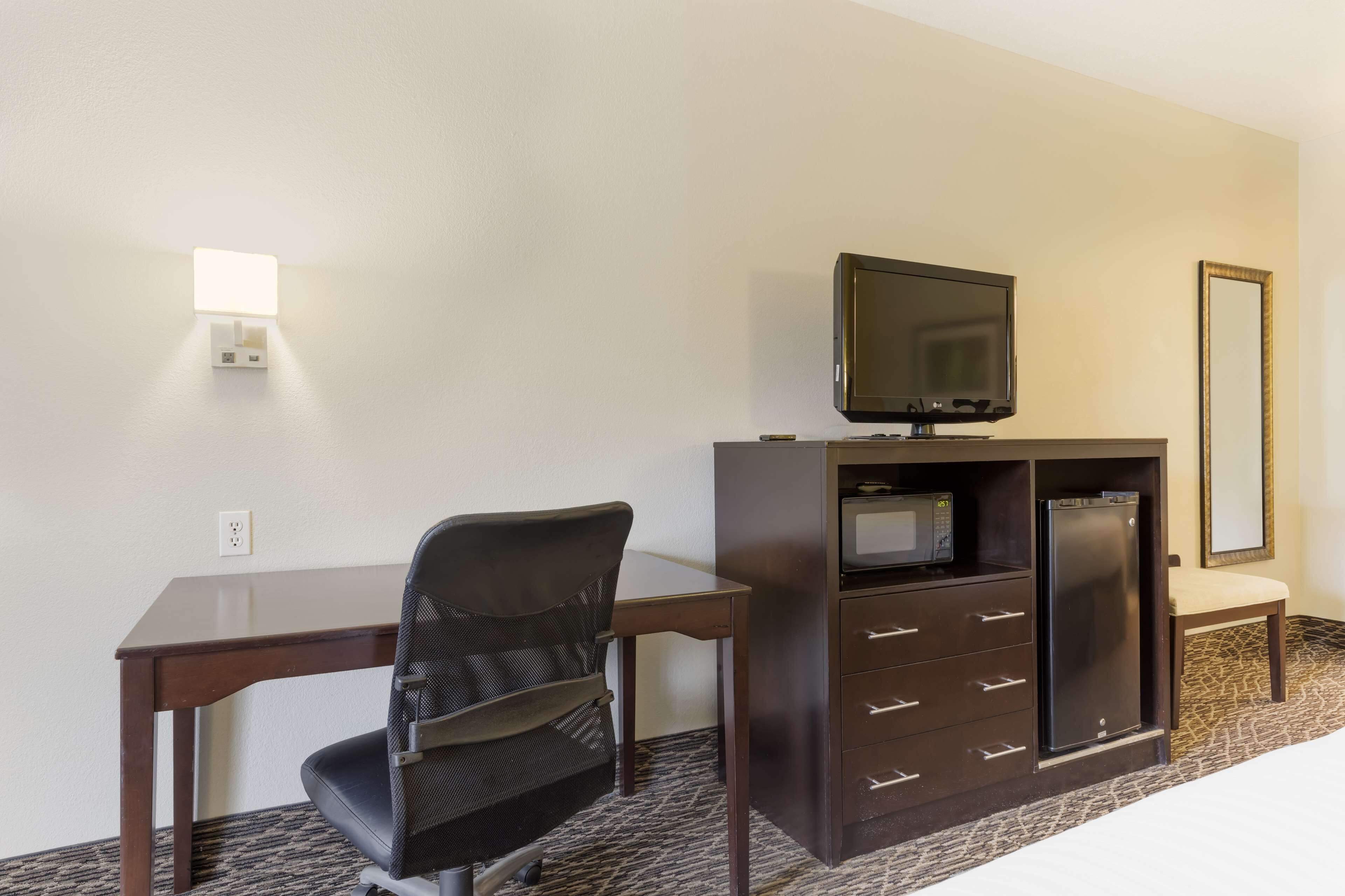 A hotel room with a desk, chair, television, minibar, and a large mirror.