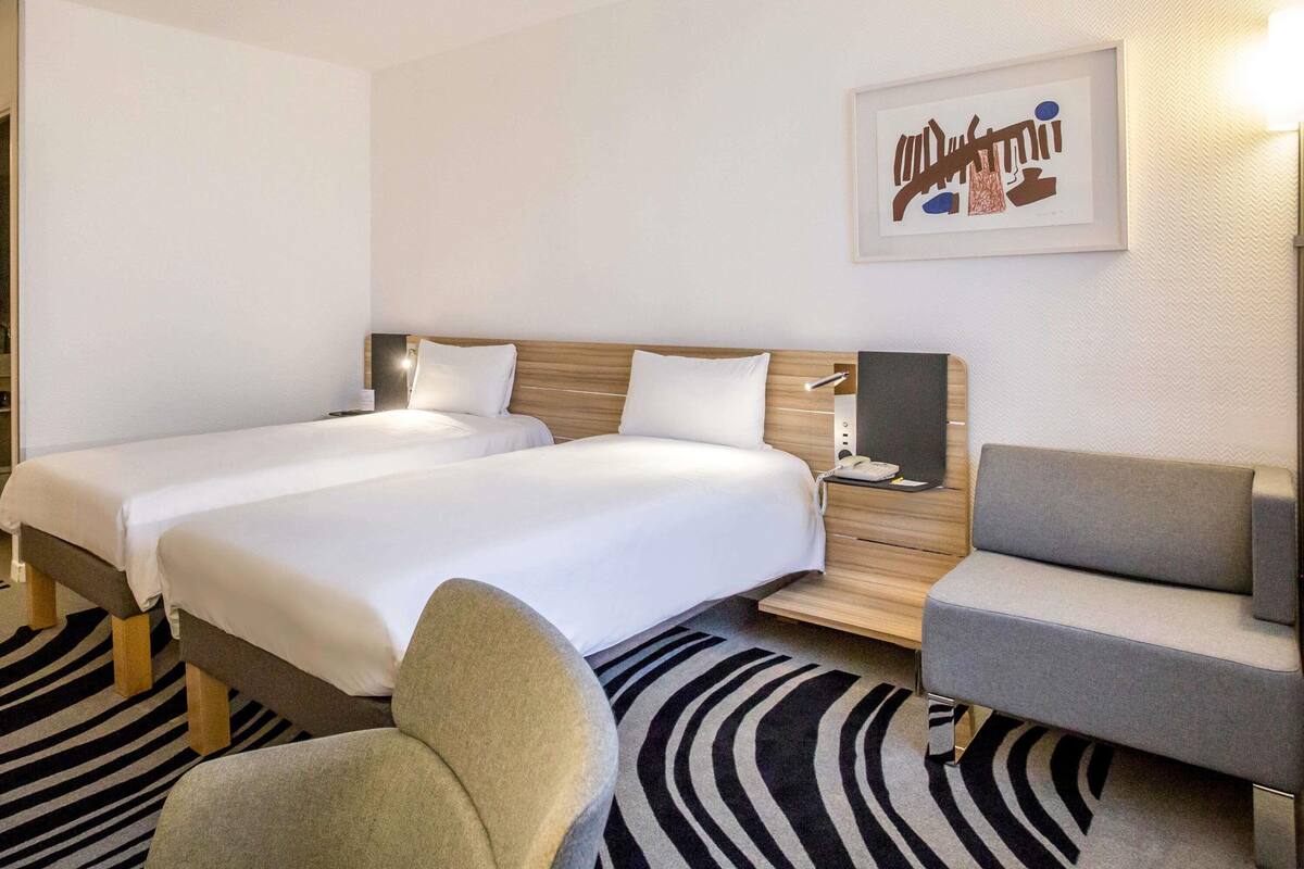 A hotel room with two beds, a desk, a chair, and a wall-mounted phone.
