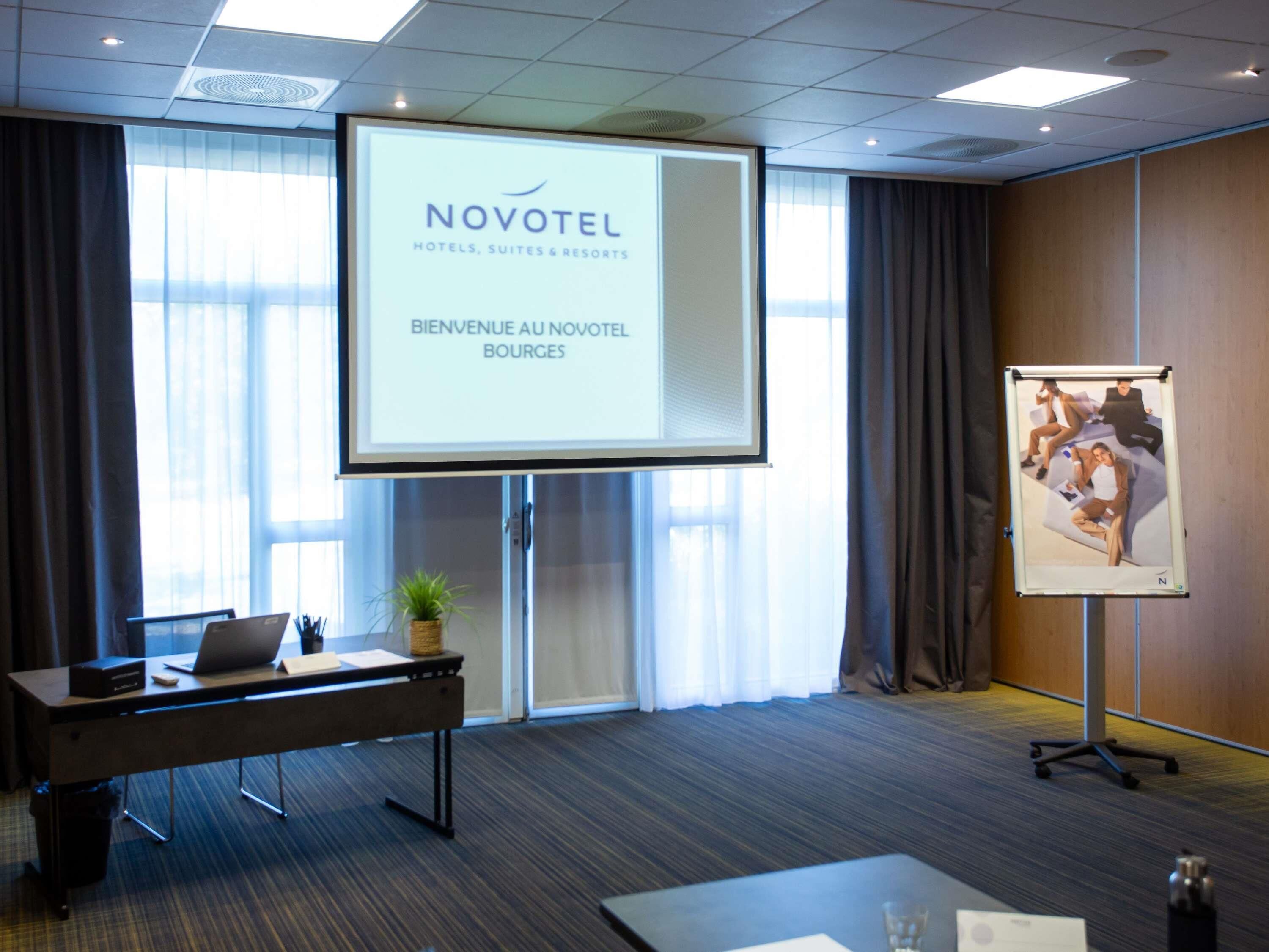 A conference room with a projection screen displaying 'NOVOTEL' and 'BIENVENUE AU NOVOTEL BOURGES'. There's a desk with a laptop, a potted plant, and a whiteboard with a photo.