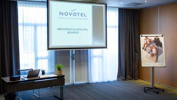 A conference room with a projection screen displaying 'NOVOTEL' and 'BIENVENUE AU NOVOTEL BOURGES'. There's a desk with a laptop, a potted plant, and a whiteboard with a photo.