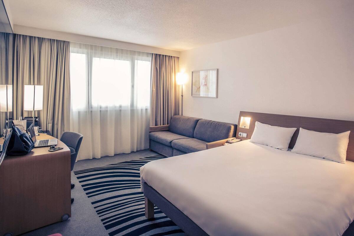 Habitación de hotel con cama, sofá, escritorio con computadora portátil, silla y ventana con cortinas.