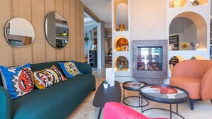 A modern living room with a green sofa, a red armchair, and a fireplace. There are two round tables and a large mirror on the wall.