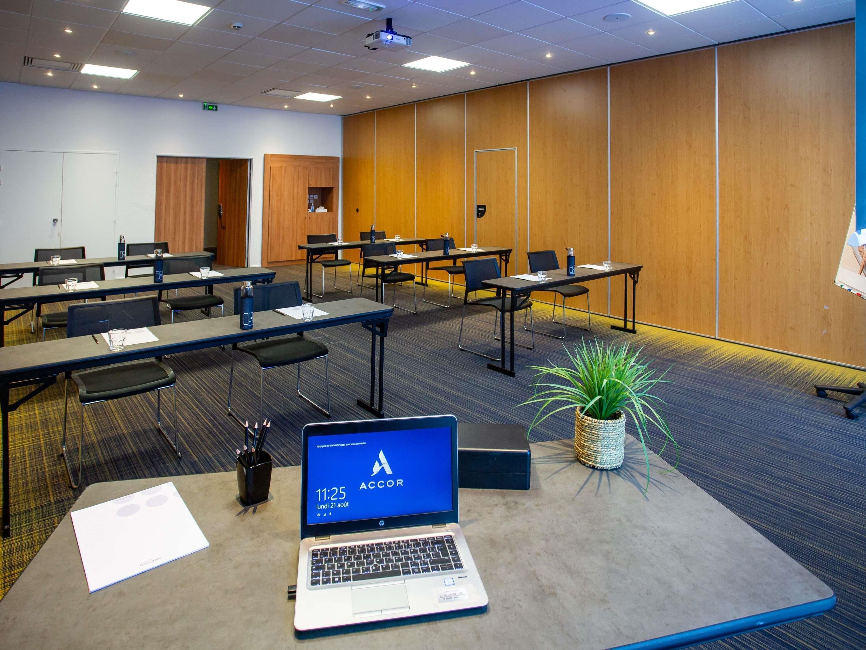 Ein Konferenzraum mit mehreren Tischen, Stühlen, einem Laptop und einem Blumentopf.