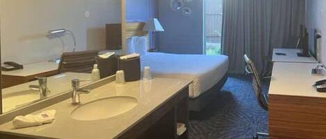 A hotel room with a large bed, a desk, a vanity with a mirror, and a bathroom with a sink and toilet.