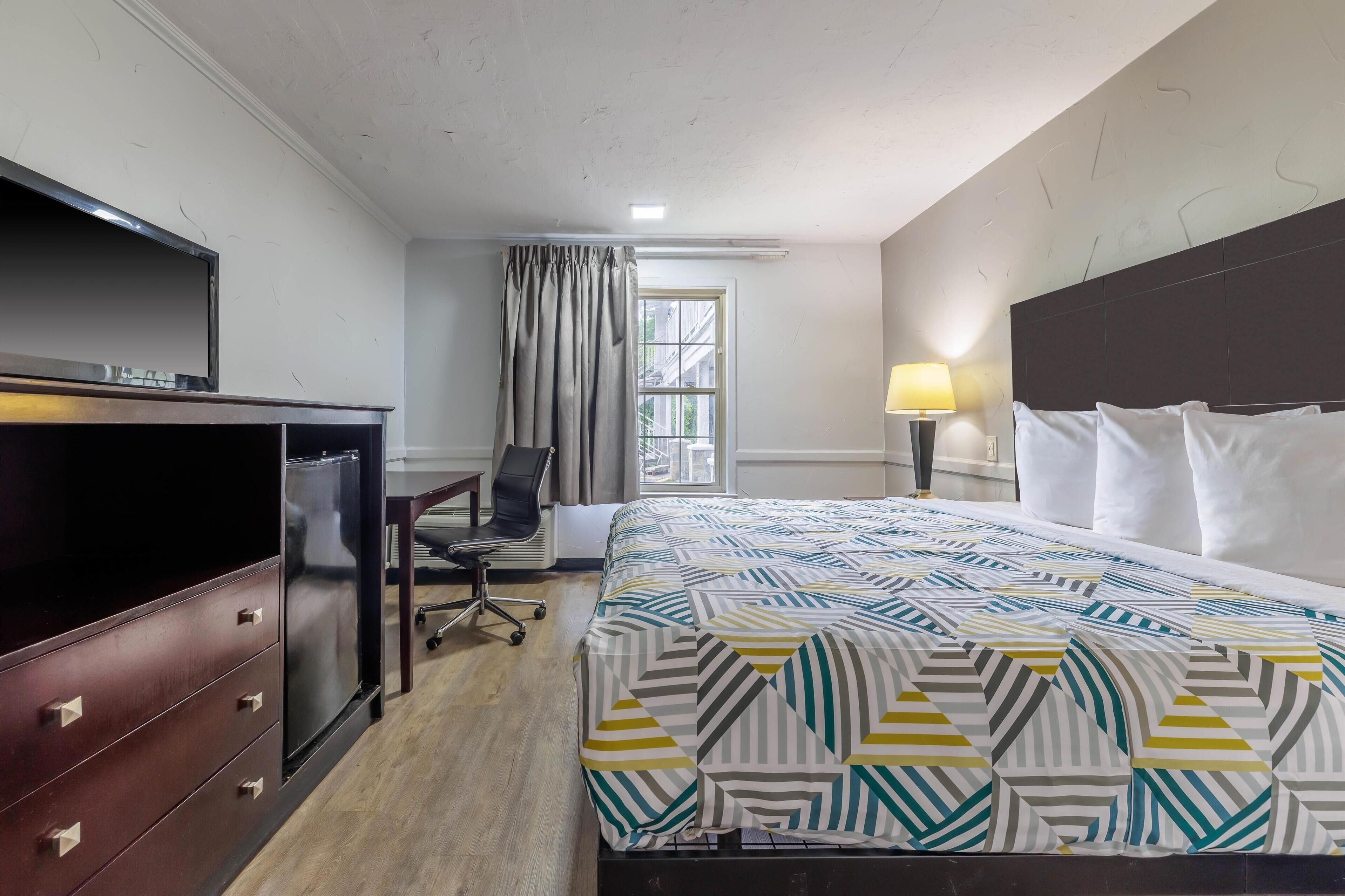 Ein Hotelzimmer mit Bett, Schreibtisch, Stuhl, Fernseher und einem fenster mit Vorhängen.