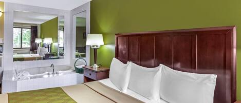 Habitación de hotel con una cama grande, cabecera de madera, pared verde, un espejo amplio, mesita de noche con lámpara y baño con bañera y lavamanos.