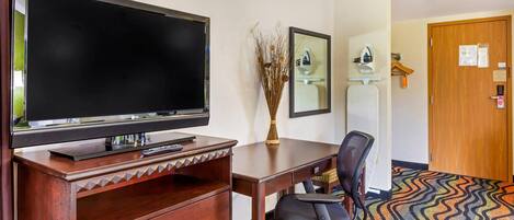 A hotel room with a flat-screen TV, a wooden desk, a chair, a vase with dried plants, a mirror, and a door with a wooden panel.