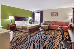 A hotel room with a bed, a desk, a chair, a sofa, a window with curtains, and a wall art piece.