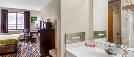 Ein Hotelzimmer mit Bett, Schreibtisch, Stuhl und einem großzügigen Badezimmer mit Waschbecken und Spiegel.