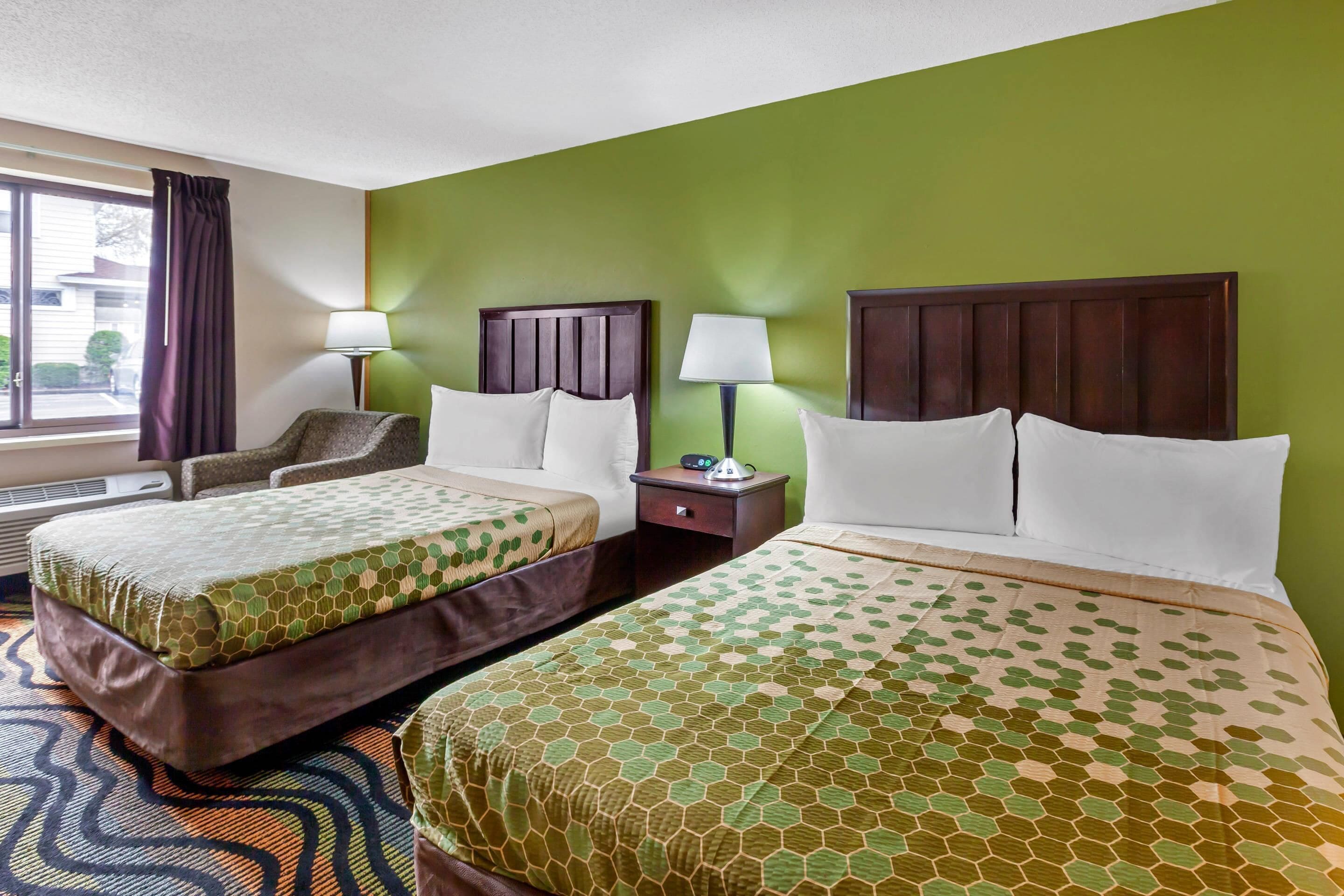 Habitación de hotel con dos camas, una silla, una mesita de noche con lámpara y una ventana con cortinas.