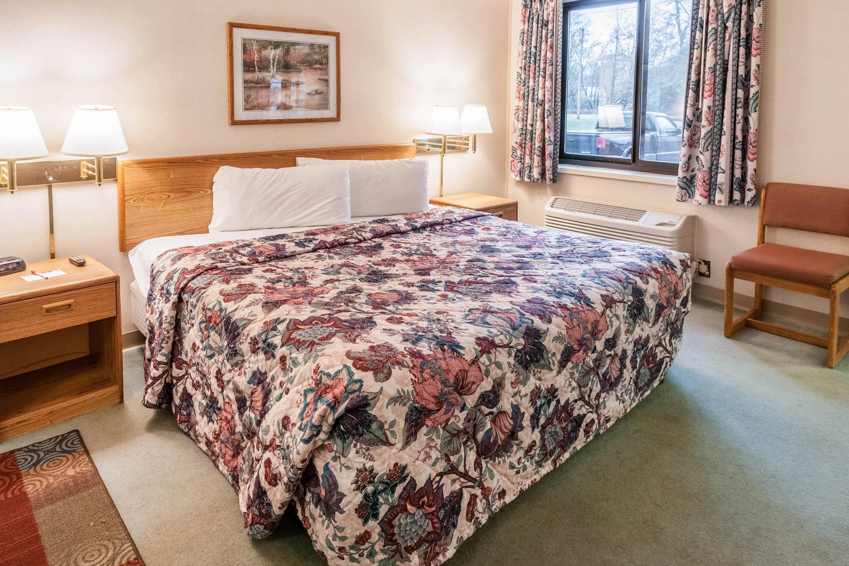 A hotel room with a large bed, two bedside lamps, a nightstand, a chair, a window with curtains, and a radiator.