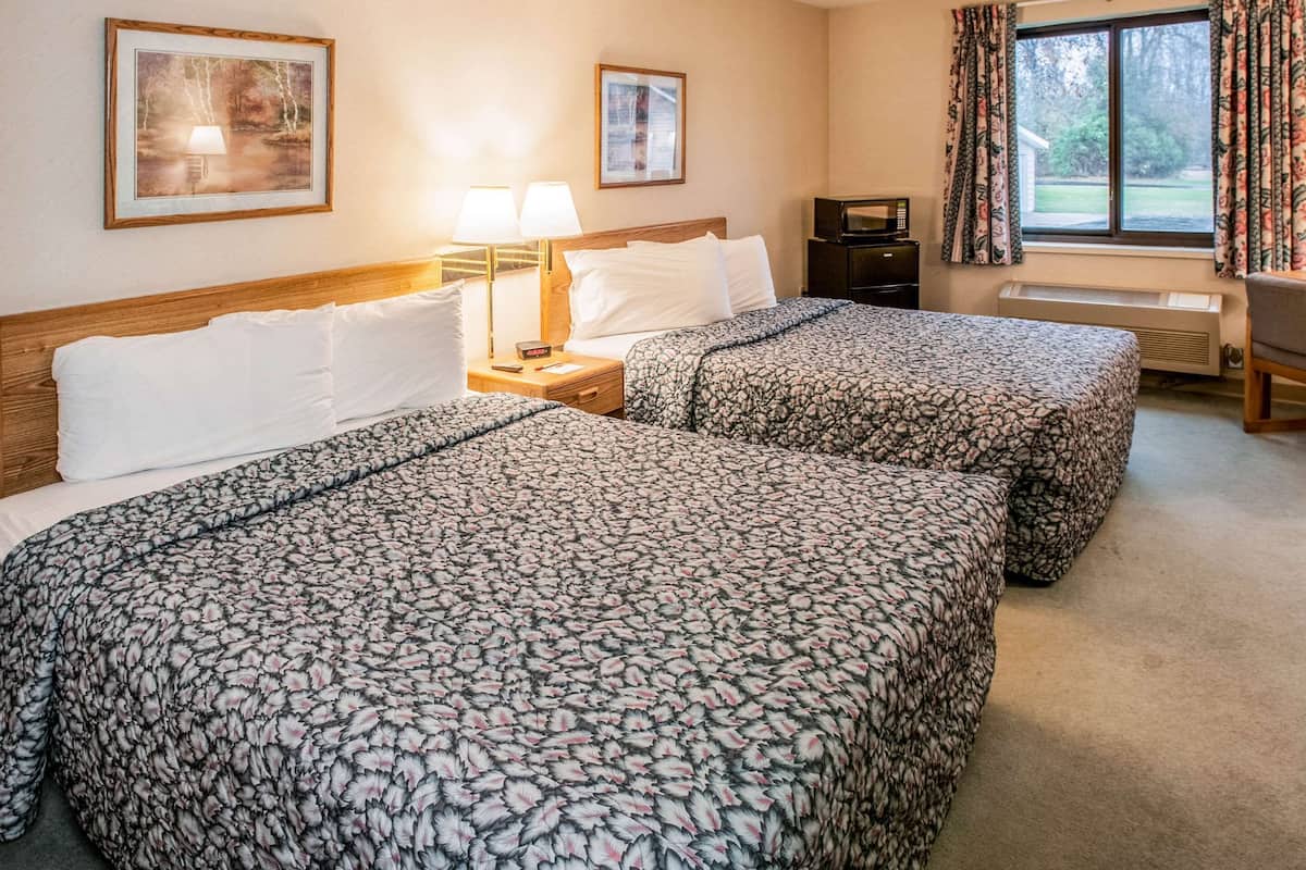 A hotel room with two beds, a nightstand with lamps, a desk with a computer, and a window with curtains.
