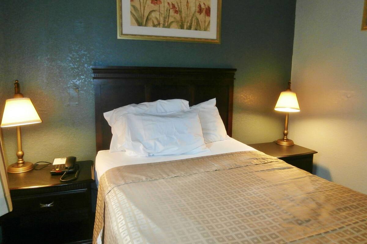 Een net opgemaakt bed met witte lakens, twee bedlampen, een telefoon en een lijst met bloemkunst op de muur.