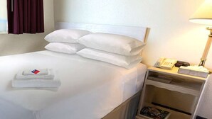 Una cama bien hecha con sábanas y almohadones blancos, una mesita de noche con una lámpara y un teléfono, y una ventana con cortinas.