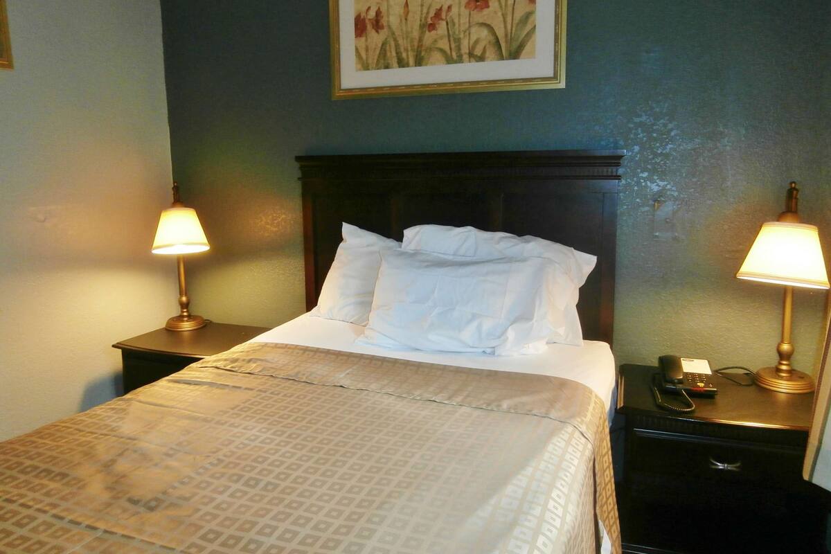 Een net opgemaakt bed met witte lakens, twee bedlampen, een telefoon en een lijst met bloemistisch kunstwerk aan de muur.