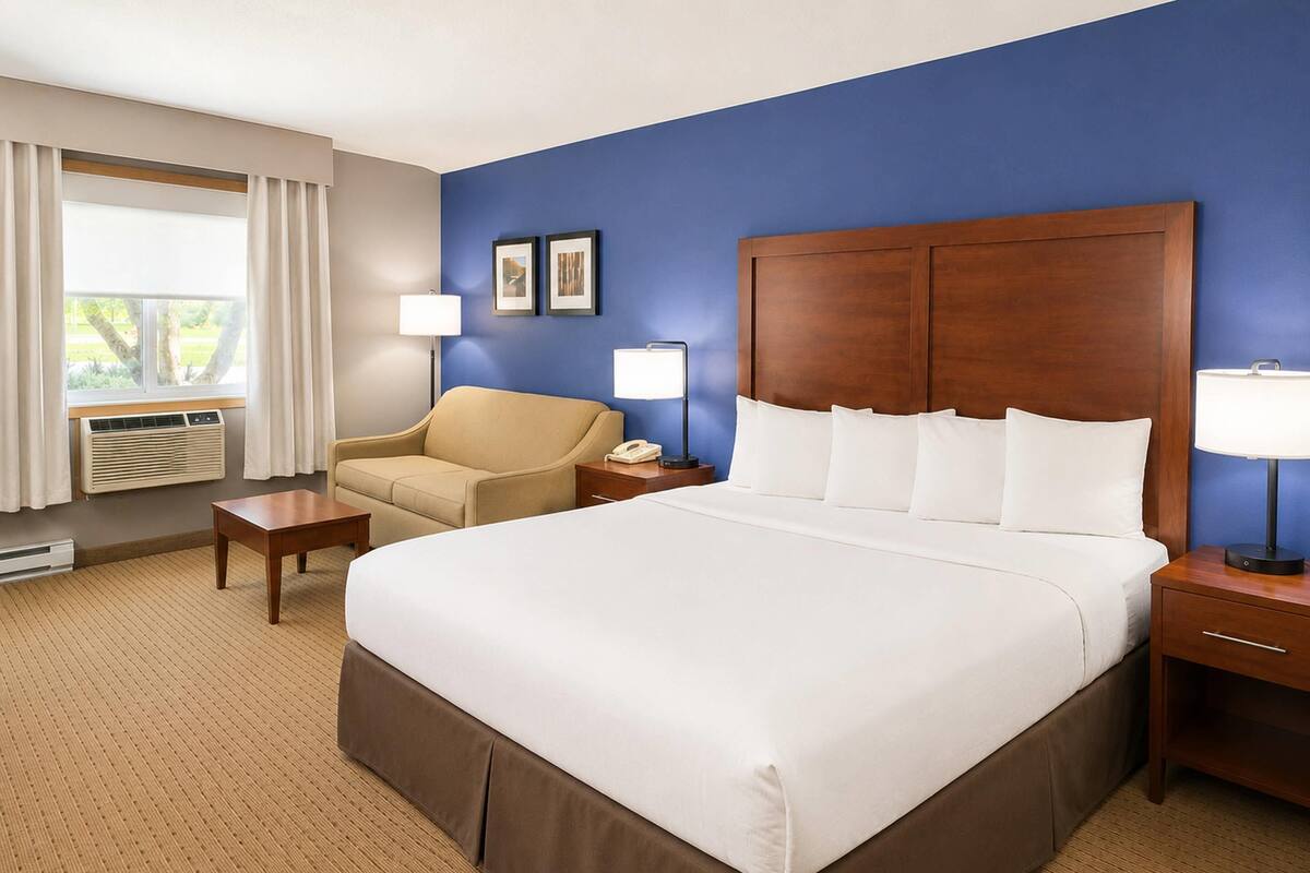 A hotel room with a large bed, a chair, a bedside table, a lamp, and a window with curtains.
