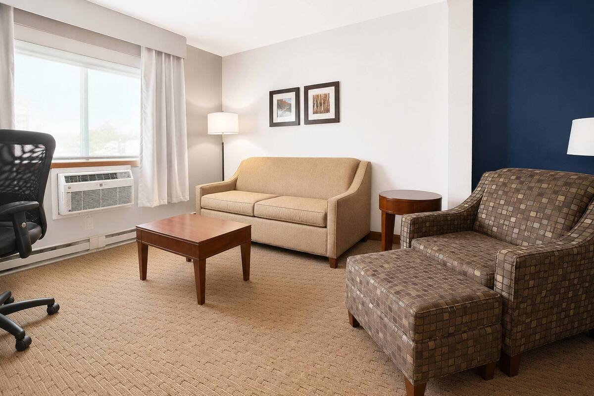 A hotel room with a beige sofa, a patterned armchair, a wooden coffee table, a black office chair, and a standing lamp.