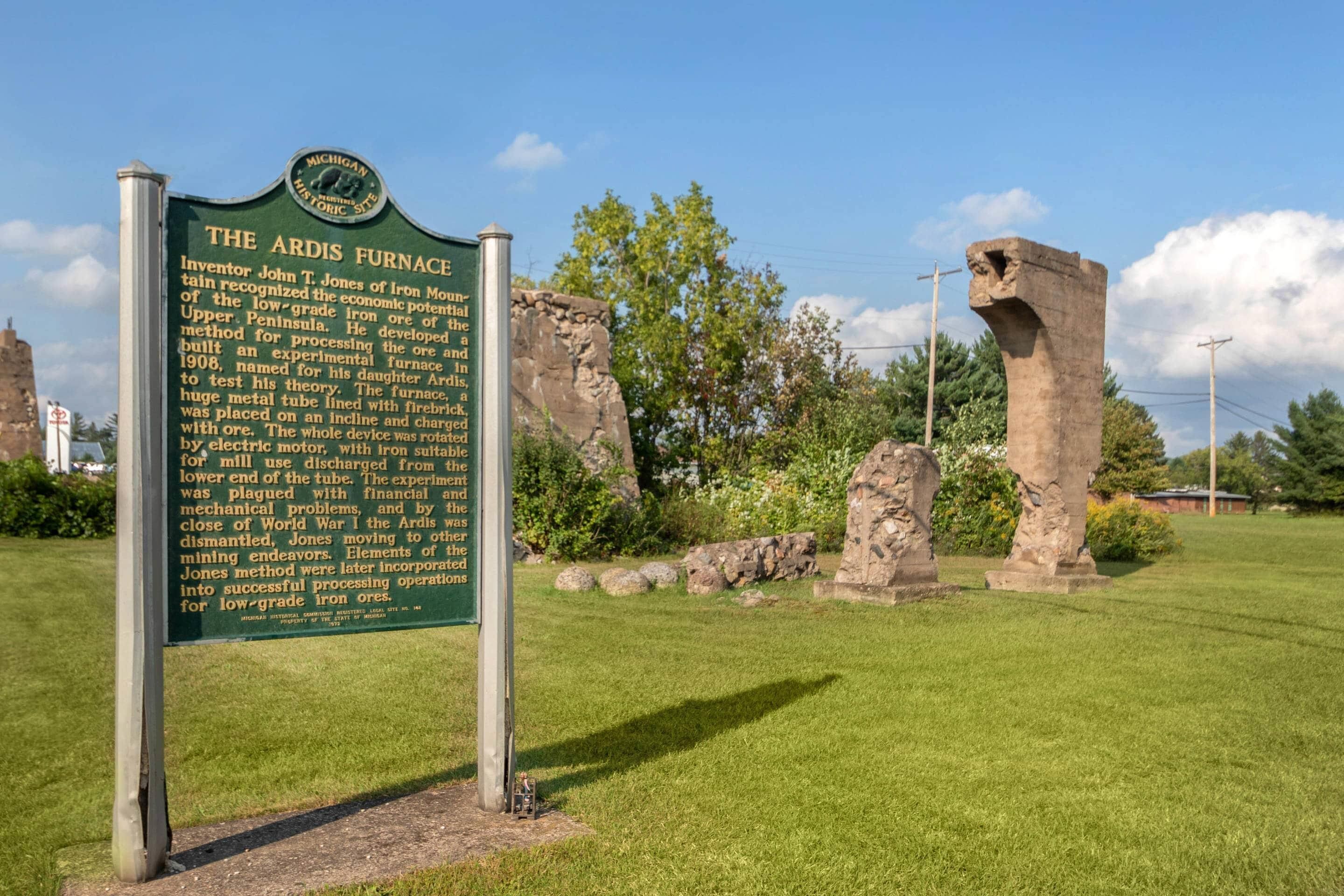 Placa de indicador histórico com texto sobre o Ardis Furnace, cercada por uma área gramada e estruturas de pedra.