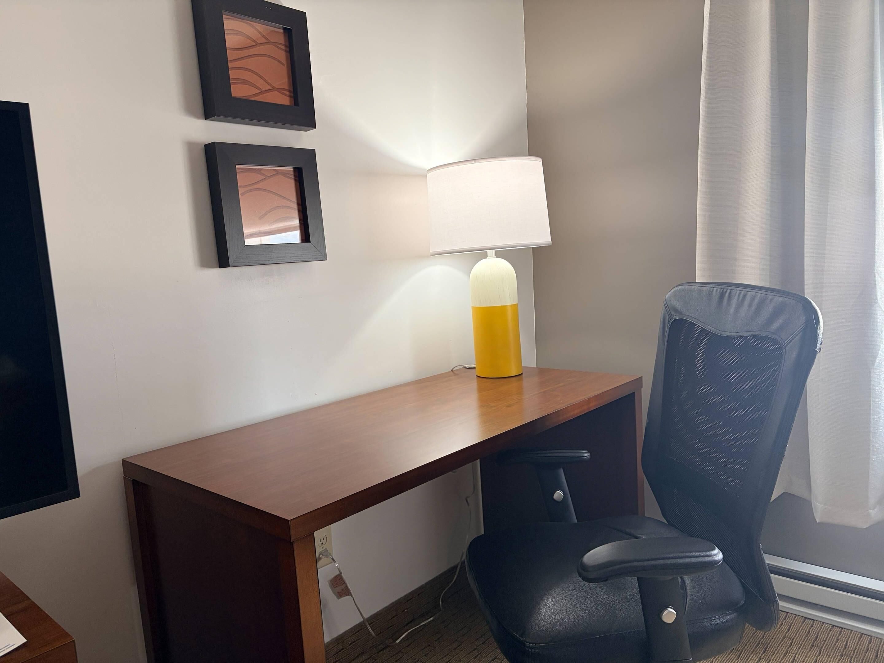 Een hotelkamer met een houten bureau, een zwarte bureaustoel, een lamp en twee ingelijste foto's aan de muur.