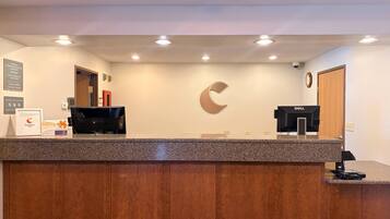 A hotel reception desk with a wooden panel design, a Dell computer, a large C-shaped logo on the wall, and a clock.