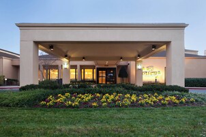 Entrada de hotel con galería techada, zona de recepción y jardín ajardinado con flores amarillas.