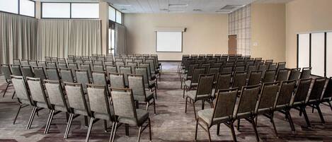 Un salón de conferencias con filas de sillas, una pantalla de proyector y un ventanal grande.
