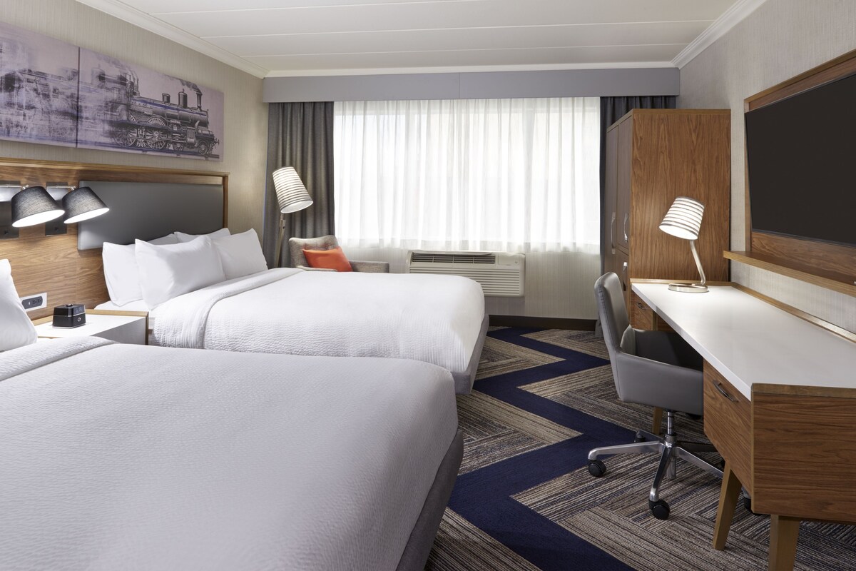 A hotel room with two beds, a desk, a chair, a desk lamp, a large window with curtains, and a painting on the wall.