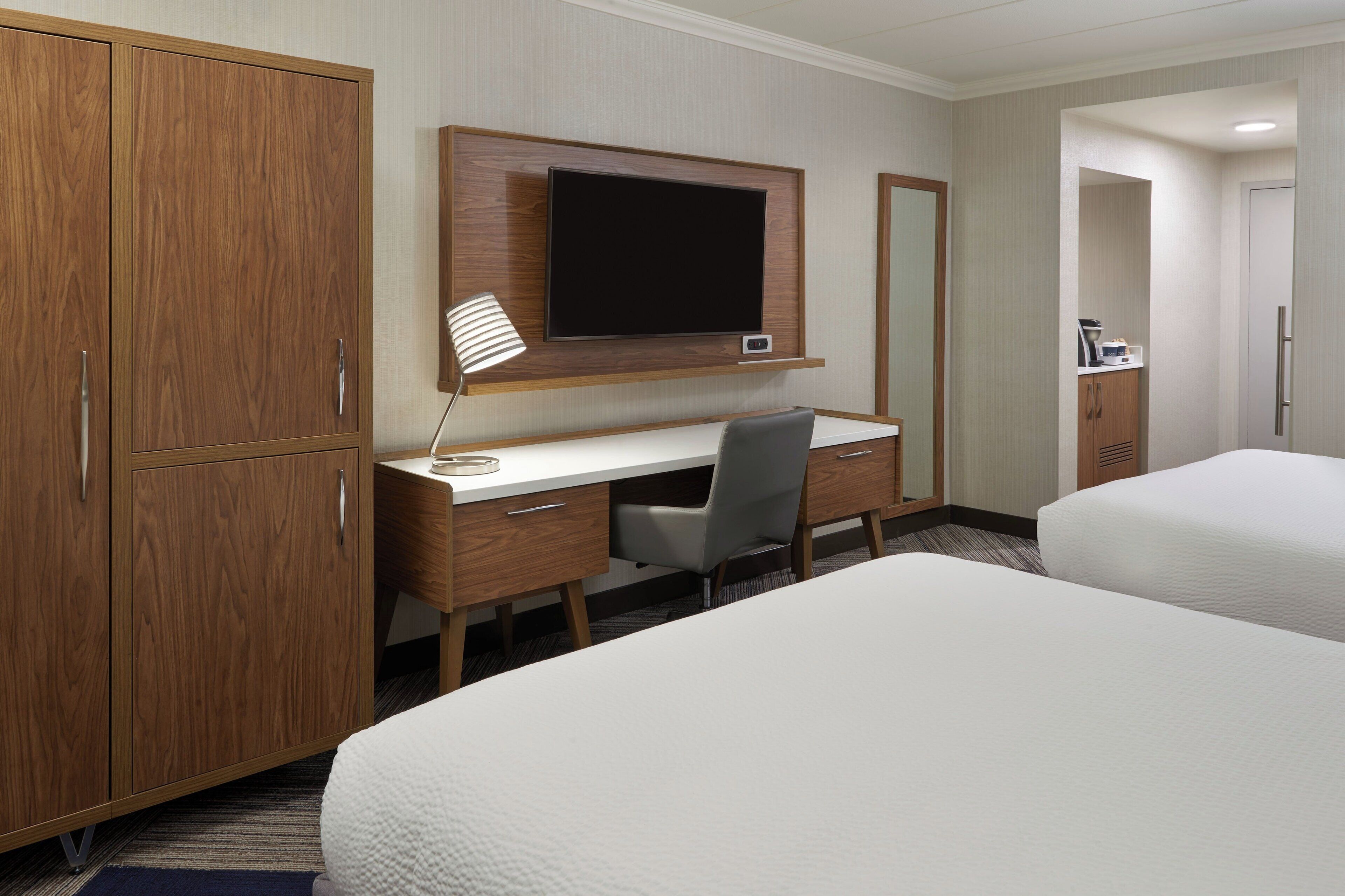 Ett hotellrum med en garderob i trä, ett skrivbord med en stol, en platt-TV och två sängar.