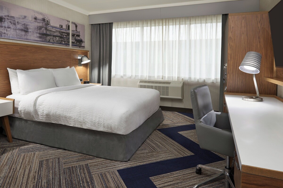 A hotel room with a large bed, a desk, a chair, a lamp, and a window with curtains.