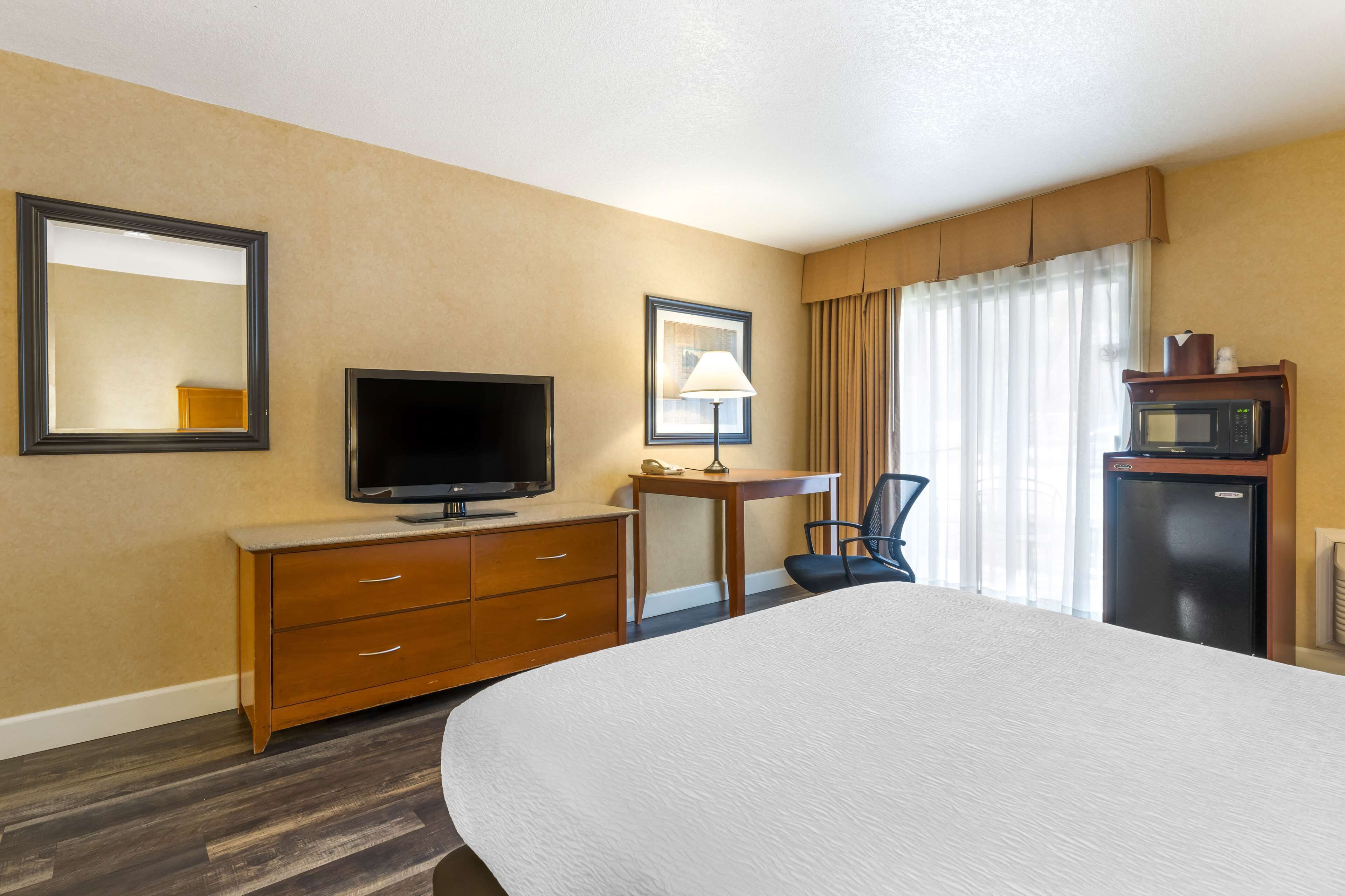 A hotel room with a large bed, a television on a wooden dresser, a desk with a lamp, a chair, and a refrigerator.