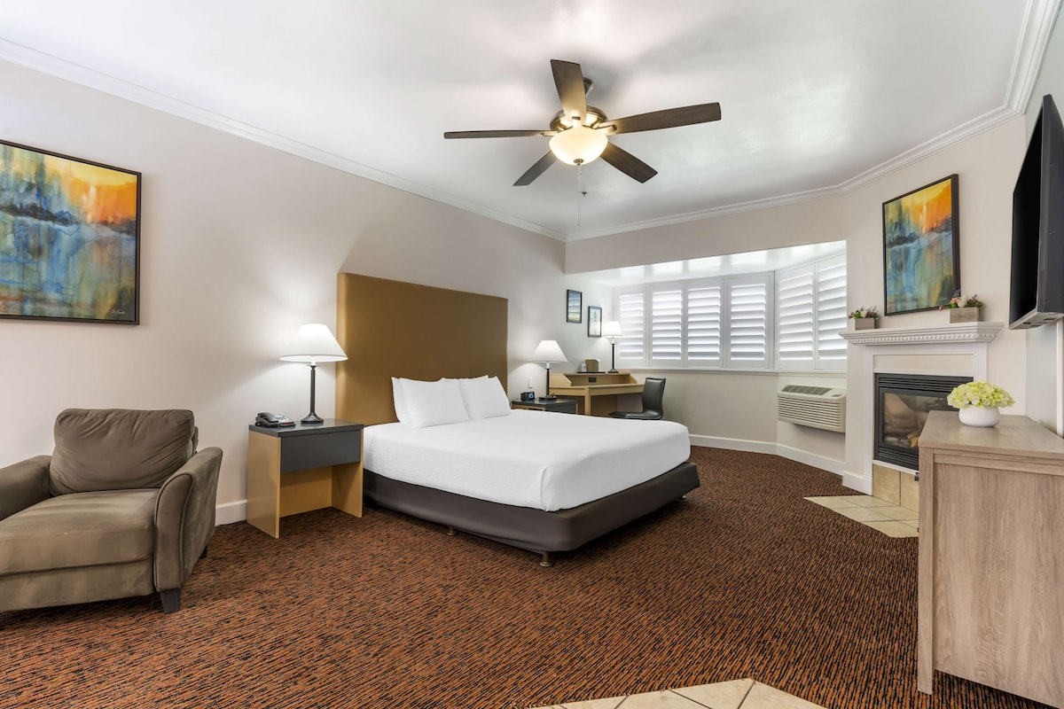 A hotel room with a large bed, a desk, a chair, a ceiling fan, and artwork on the walls.