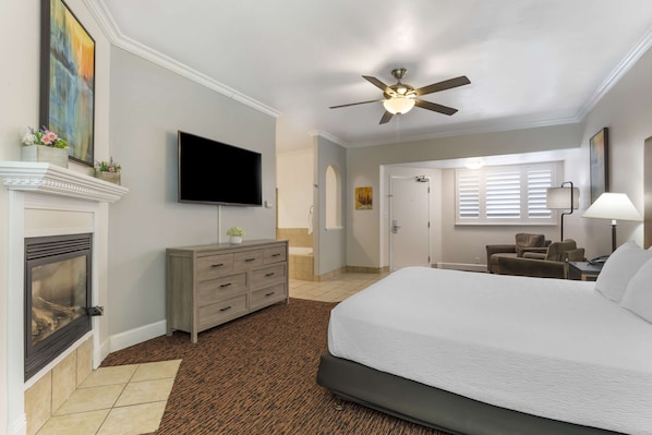 A hotel room with a large bed, a television mounted on the wall, a fireplace, a ceiling fan, and a window with blinds.