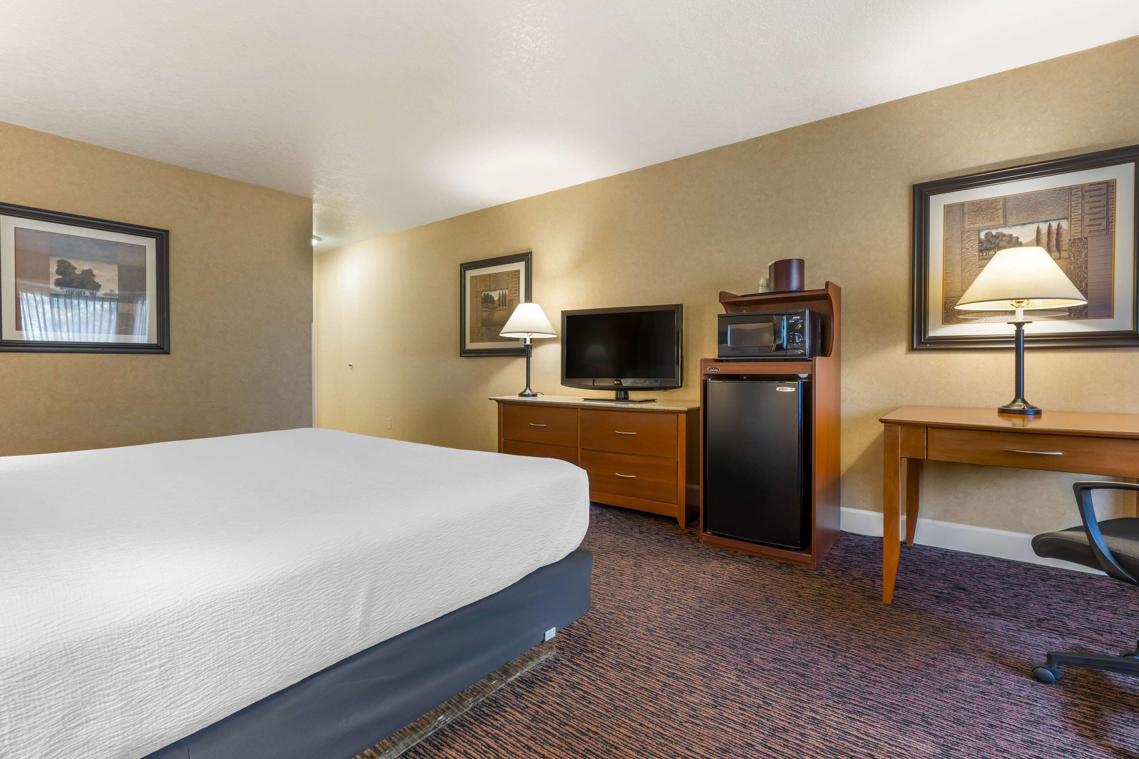 A hotel room with a bed, a desk with a computer and a lamp, a television, and a refrigerator.