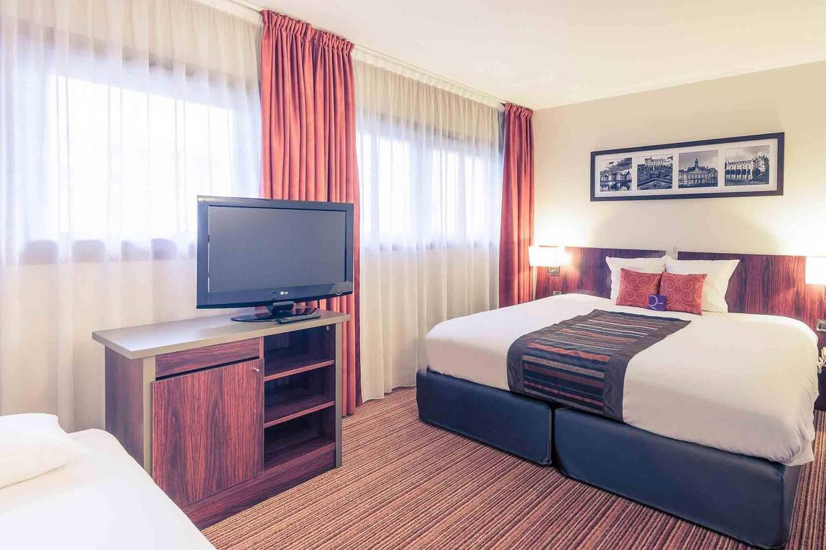 Een hotelkamer met een bed, een televisie op een standaard, een bureau met stoel, en een ingelijst schilderij aan de muur.