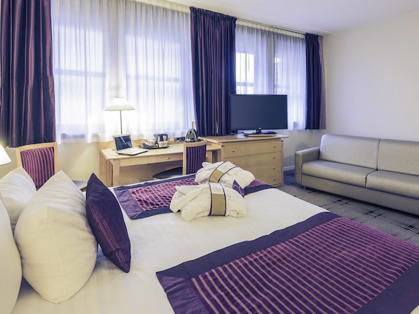 Quarto de hotel com cama, escrivaninha, televisão, sofá e cortinas.