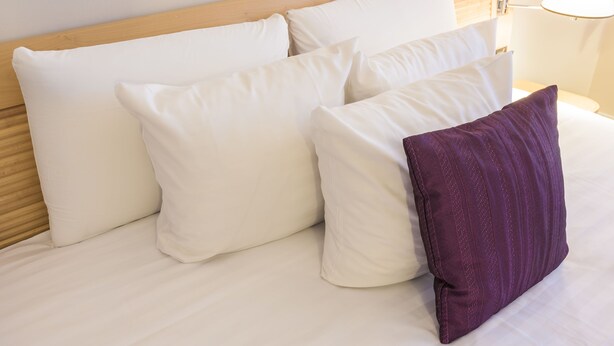 Un lit bien fait, avec des oreillers blancs et violets, une tête de lit en bois et une lampe de chevet.