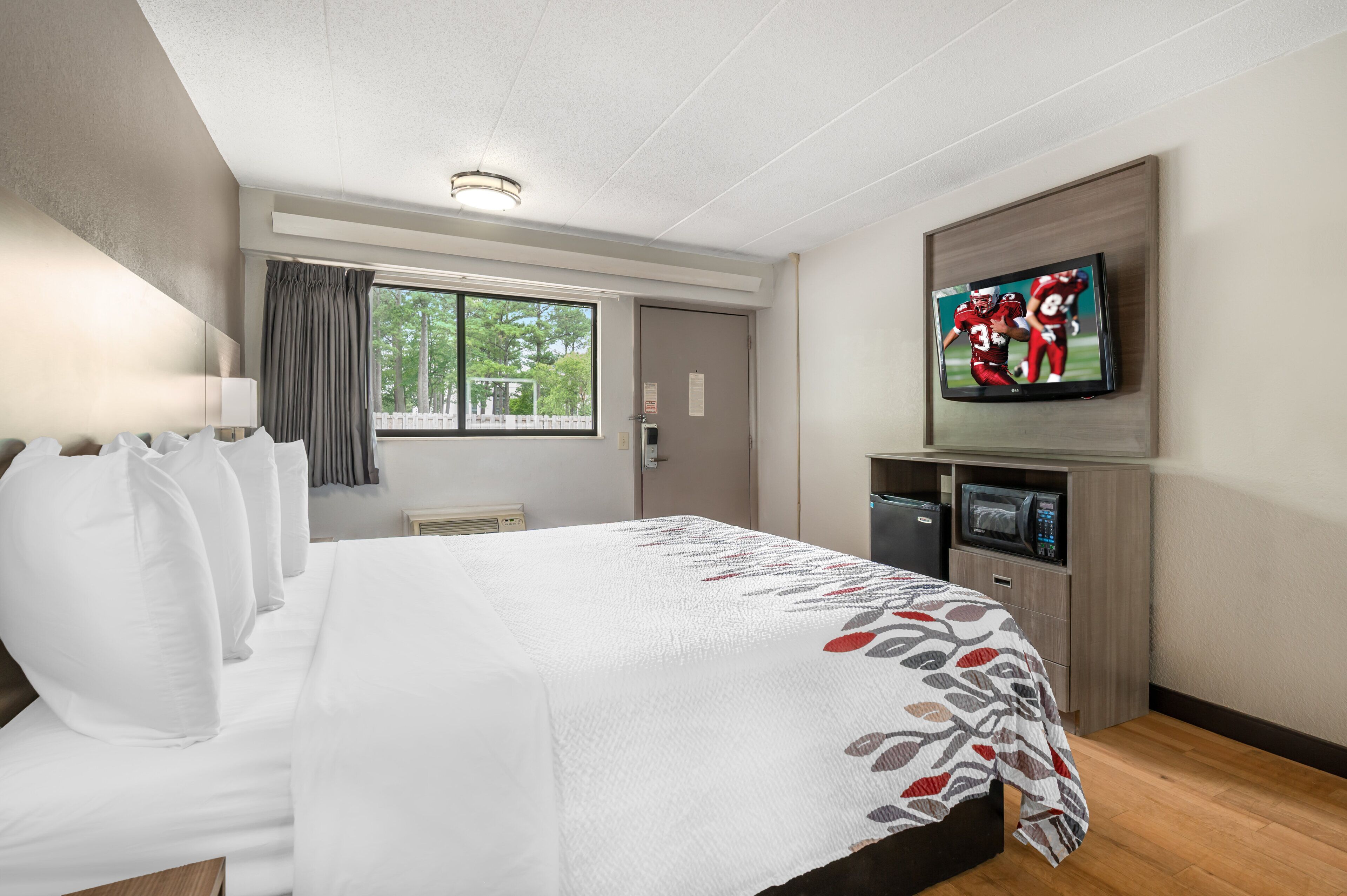 A hotel room with a large bed, a TV mounted on the wall, a window with curtains, and a microwave in the corner.