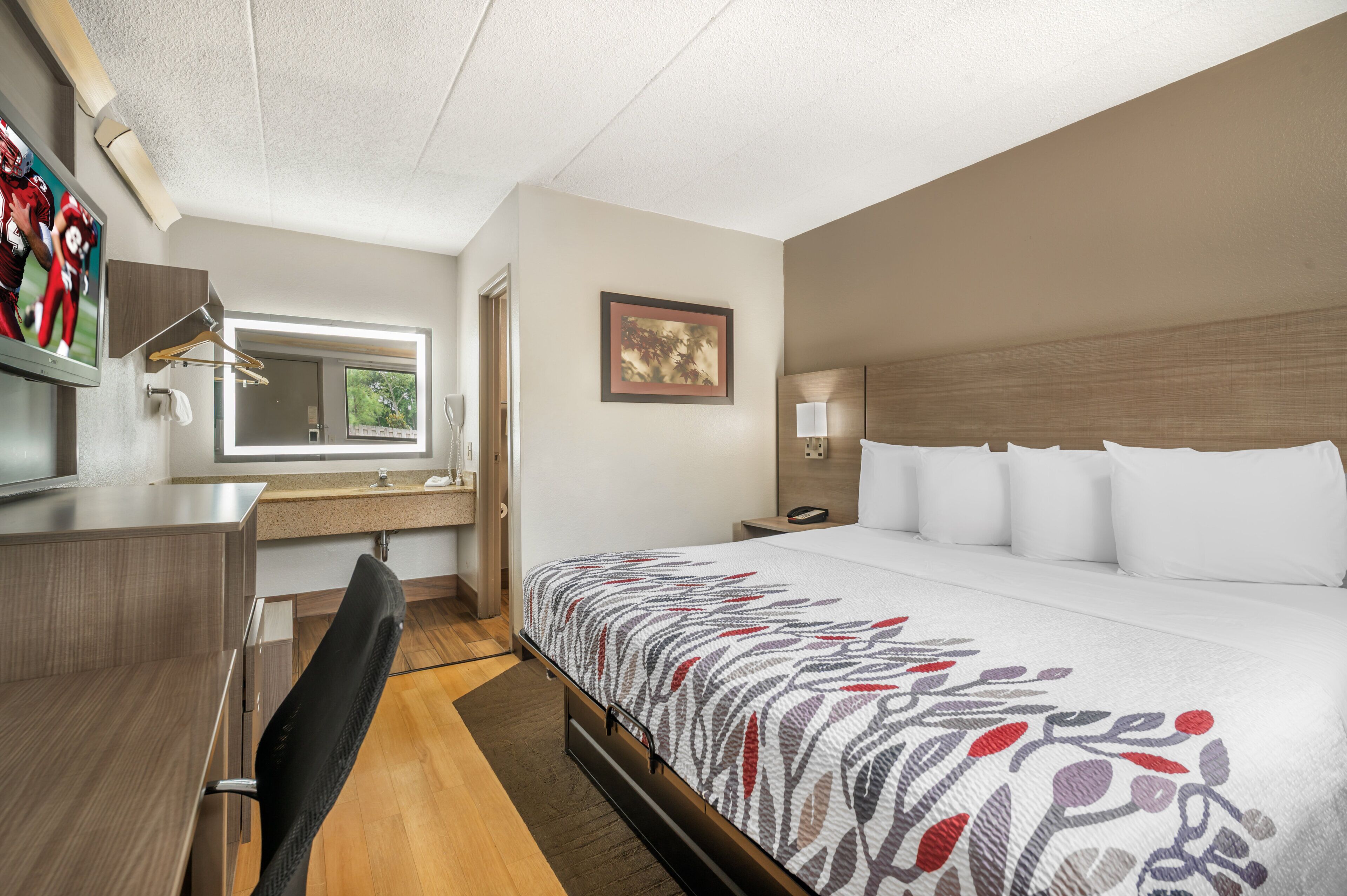 Una habitación de hotel con cama, escritorio, silla, ventana y televisor instalado en la pared.