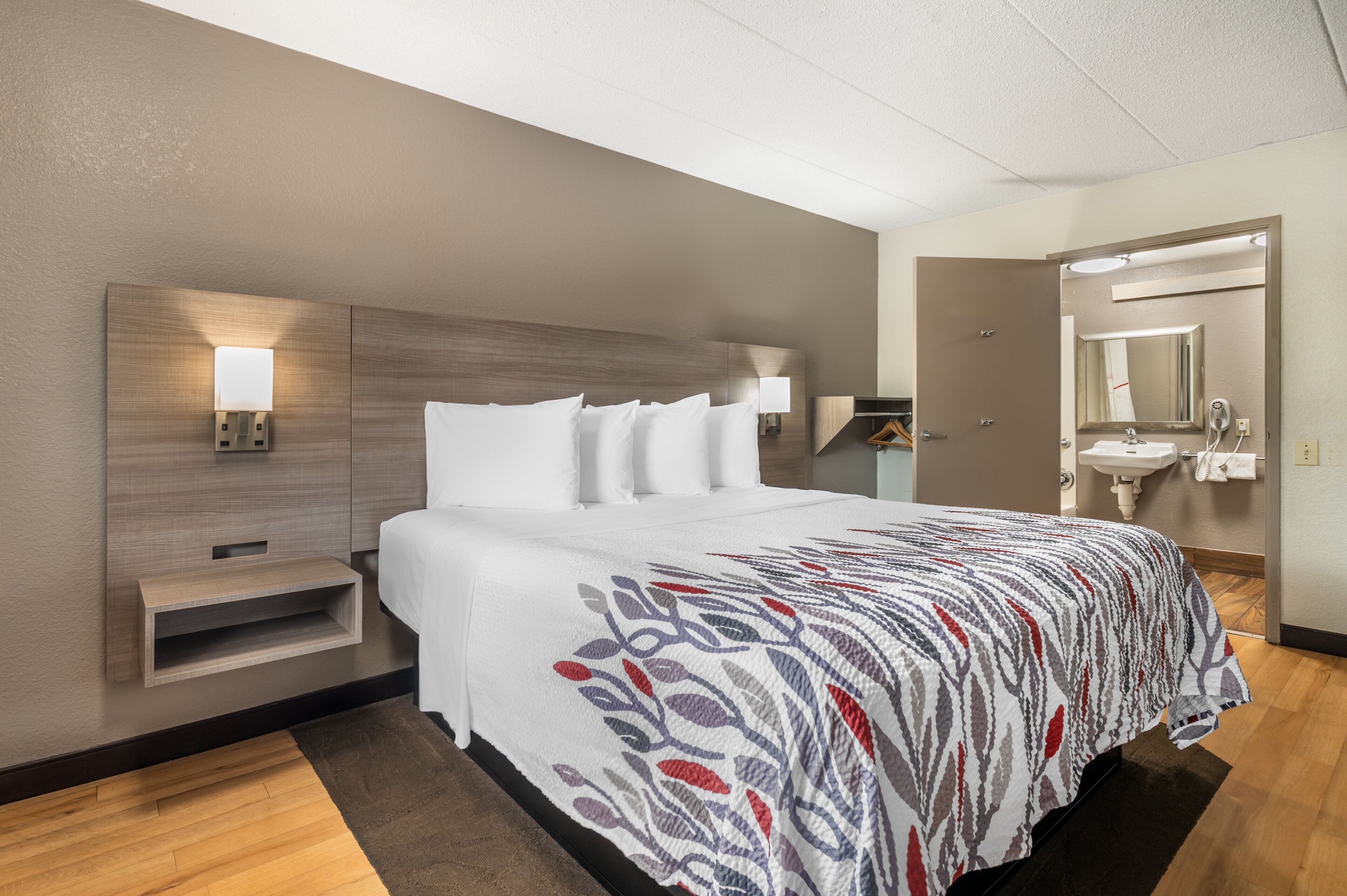 Ein Hotelzimmer mit einem Bett, Nachttischlampen, einem Nachttisch, einem Spiegel und einem Badezimmer mit Waschbecken und Spiegel.