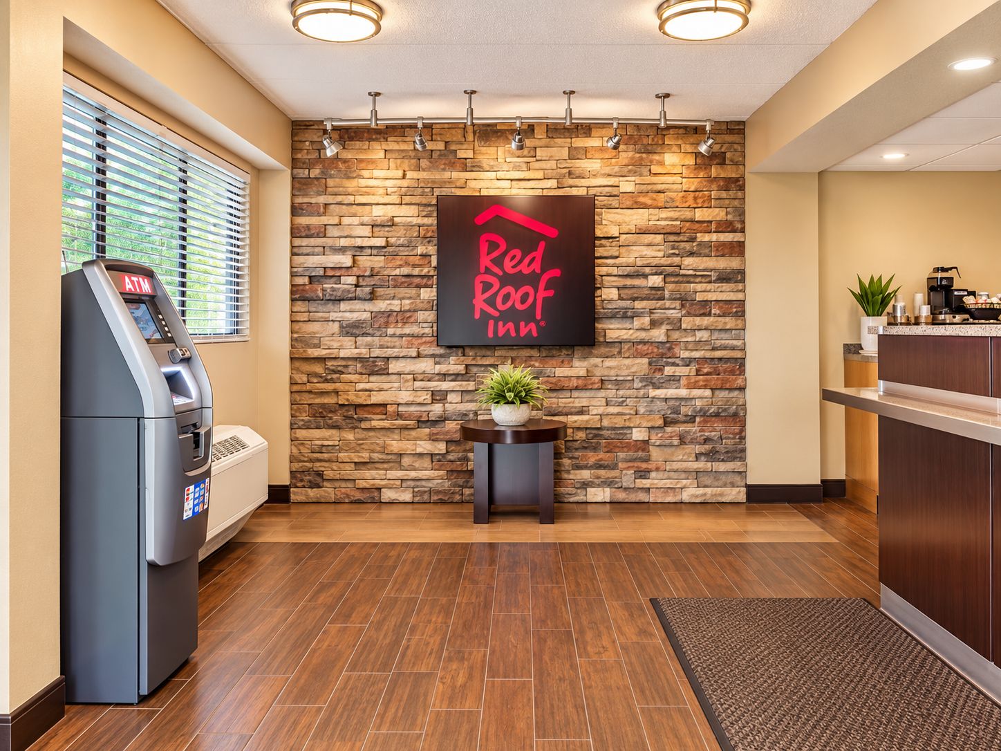 A Red Roof Inn reception area with a stone wall, an ATM, a small table with a plant, and a coffee station.