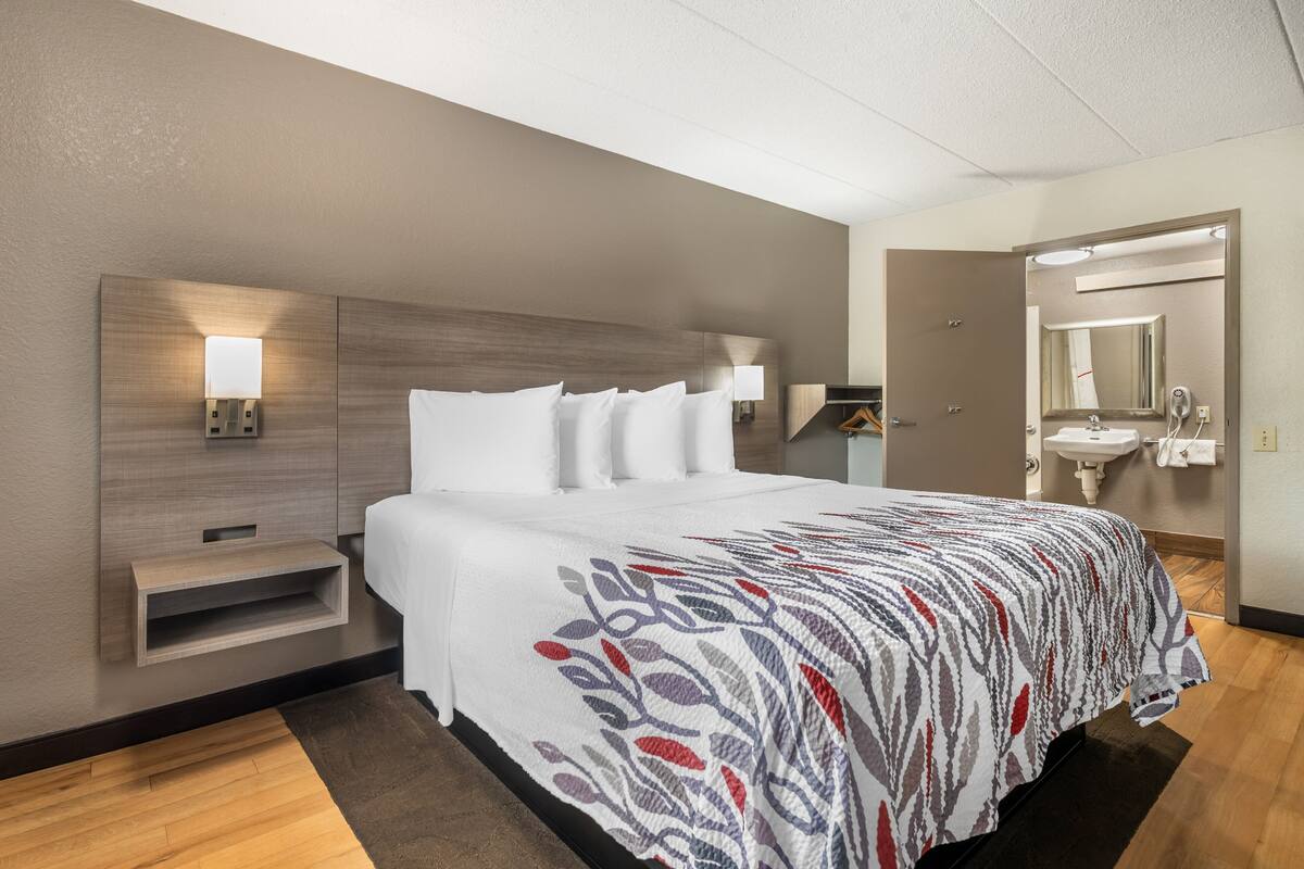 Quarto de hotel com cama, abajures, mesa de cabeceira, espelho e banheiro com pia e espelho.